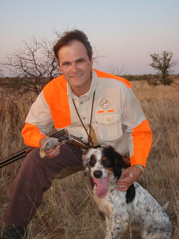 The Bob White Genome project guy kneeling down with shotgun and hunting dog. Dog sticking out his tounge and man holding dead quail bird in hand. khaki shirt orange accents on his shirt